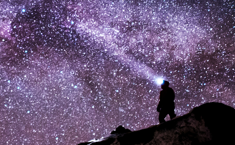 A man with a headlamp at the mountain summit under a sky full of stars