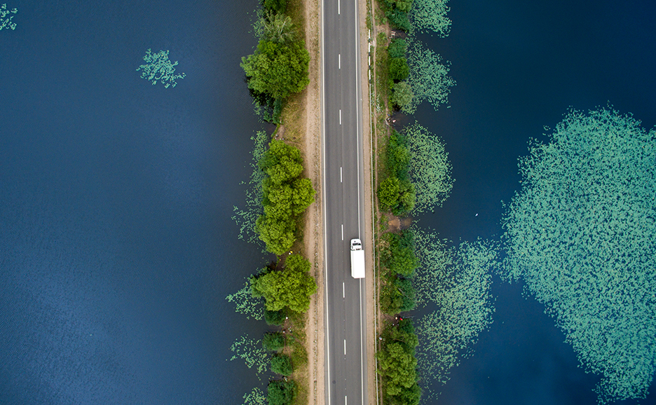 Aerial view of a road bordered by water on both sides. Lush green vegetation lines the road, and a white van is traveling down the center.