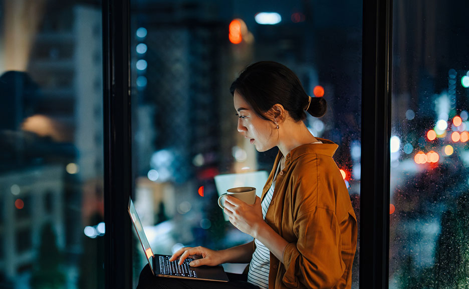 夜のライトアップされた街を背景に窓際に座ってノートパソコンを使っている女性