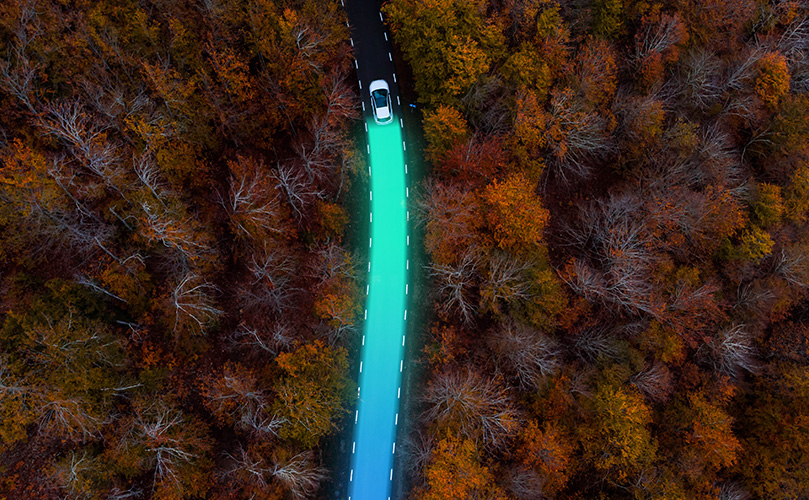 Aerial view of an electric car driving and leaving behind a sustainable trace between autumn forest
