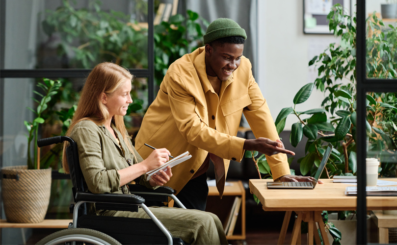Man and Woman in a wheelchair working together at an office space, smiling while looking at computer and taking notes