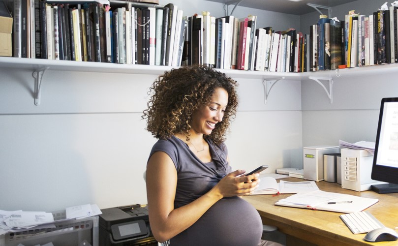  Pregnant woman smiling at the mobile phone, seated next to a desk and a shelf on the wall full of books 